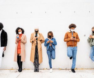 group of people distanced, in masks, leaning against a wall on their phones